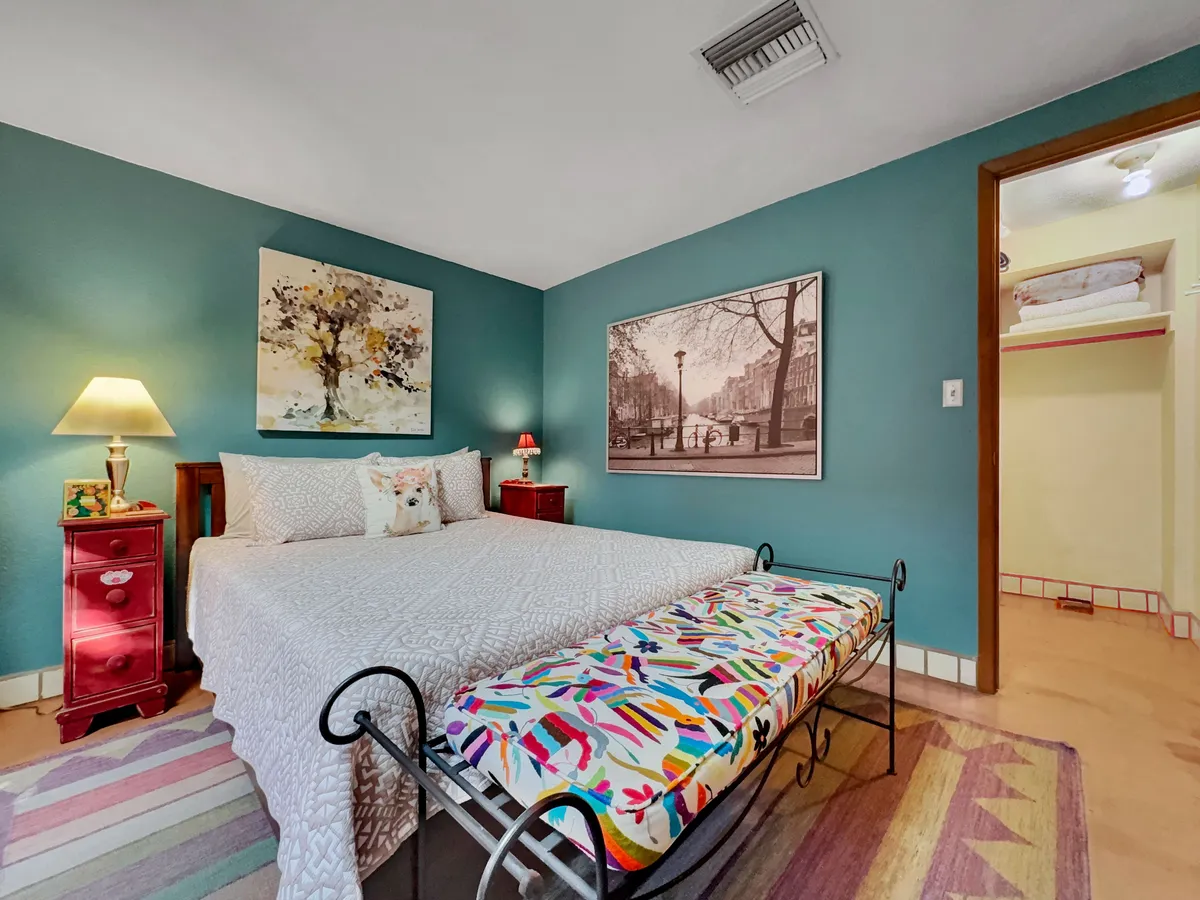A bright and welcoming sleeping area featuring a king bed dressed in a white quilt, a colorful bench at the foot of the bed, a red nightstand with a lamp, and distinctive artwork adorning the walls. Soft lighting and cheerful colors create a restful, inviting atmosphere.