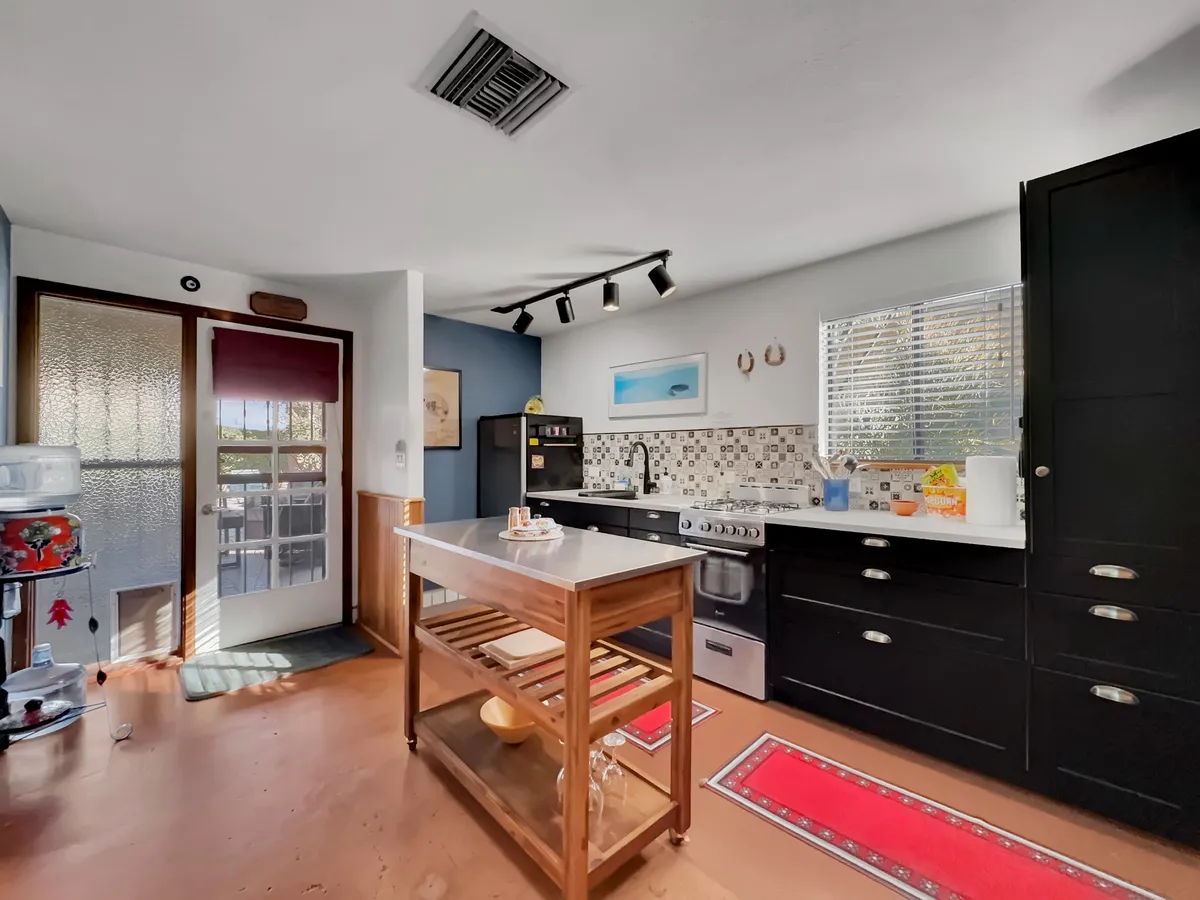 A functional kitchen scene with a kitchen island, stove, tiled back wall, and windows letting in natural light. The environment feels warm and practical, suggesting space for both cooking and gathering.