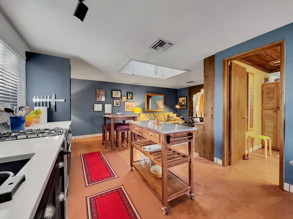 A welcoming kitchen with a central island, framed art on the walls, a comfortable yellow armchair, and a skylight that brings in plenty of natural light. The kitchen is well-equipped and aesthetically pleasing, making it suitable for both cooking and casual gatherings.