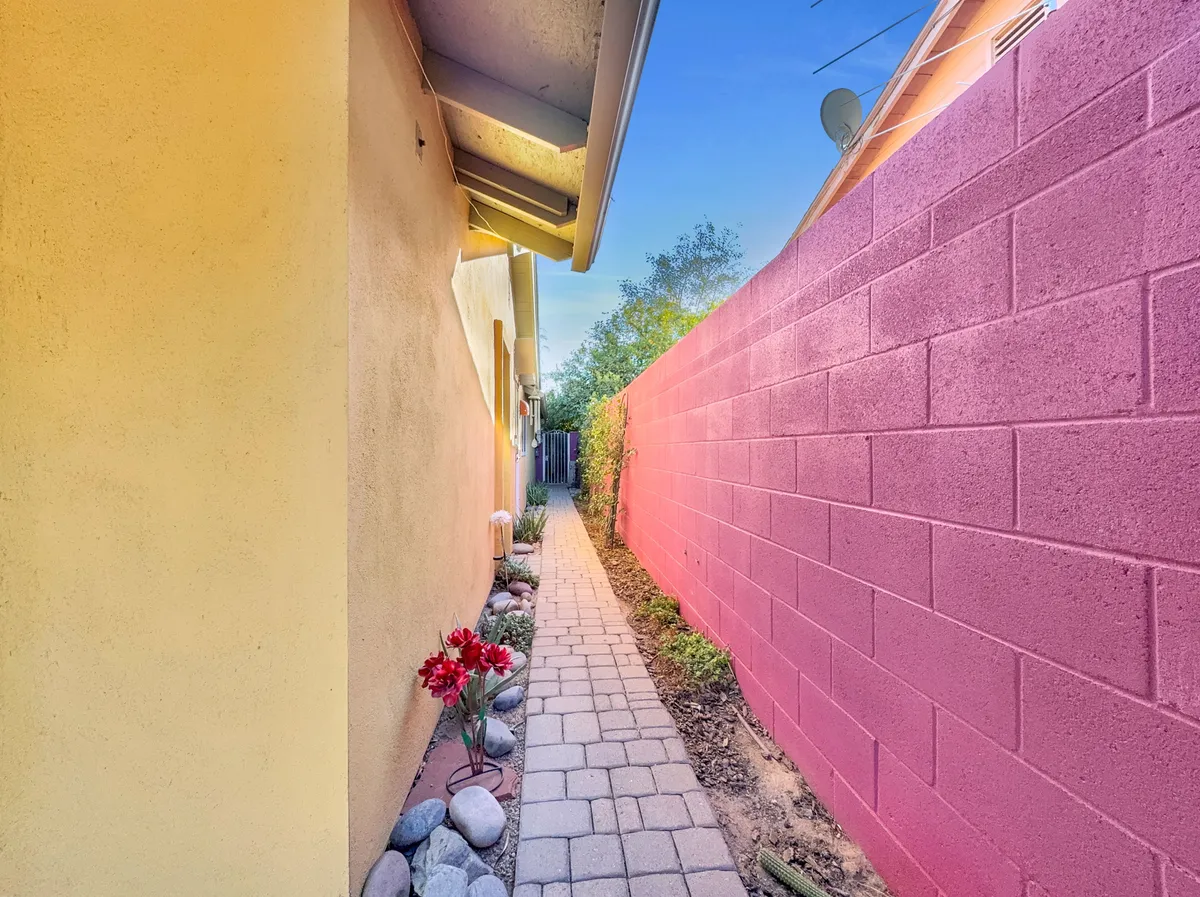A cheerful exterior scene showing a pathway flanked by vibrant yellow and pink walls, blooming flowers, and greenery. The inviting walkway leads through a garden-like setting, evoking a sense of exploration and discovery in the surrounding neighborhood.