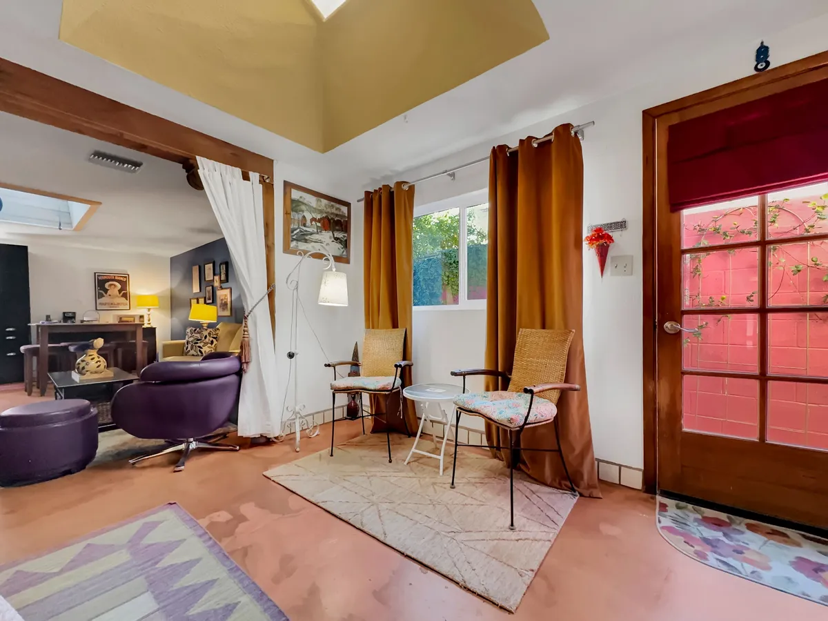 A bright and airy interior scene featuring two comfortable chairs and a small white table by a window. Natural light pours in, creating a welcoming, tranquil atmosphere. The view through the door connects the inside to the outdoors, while the setting suggests a space for relaxation or quiet reflection.