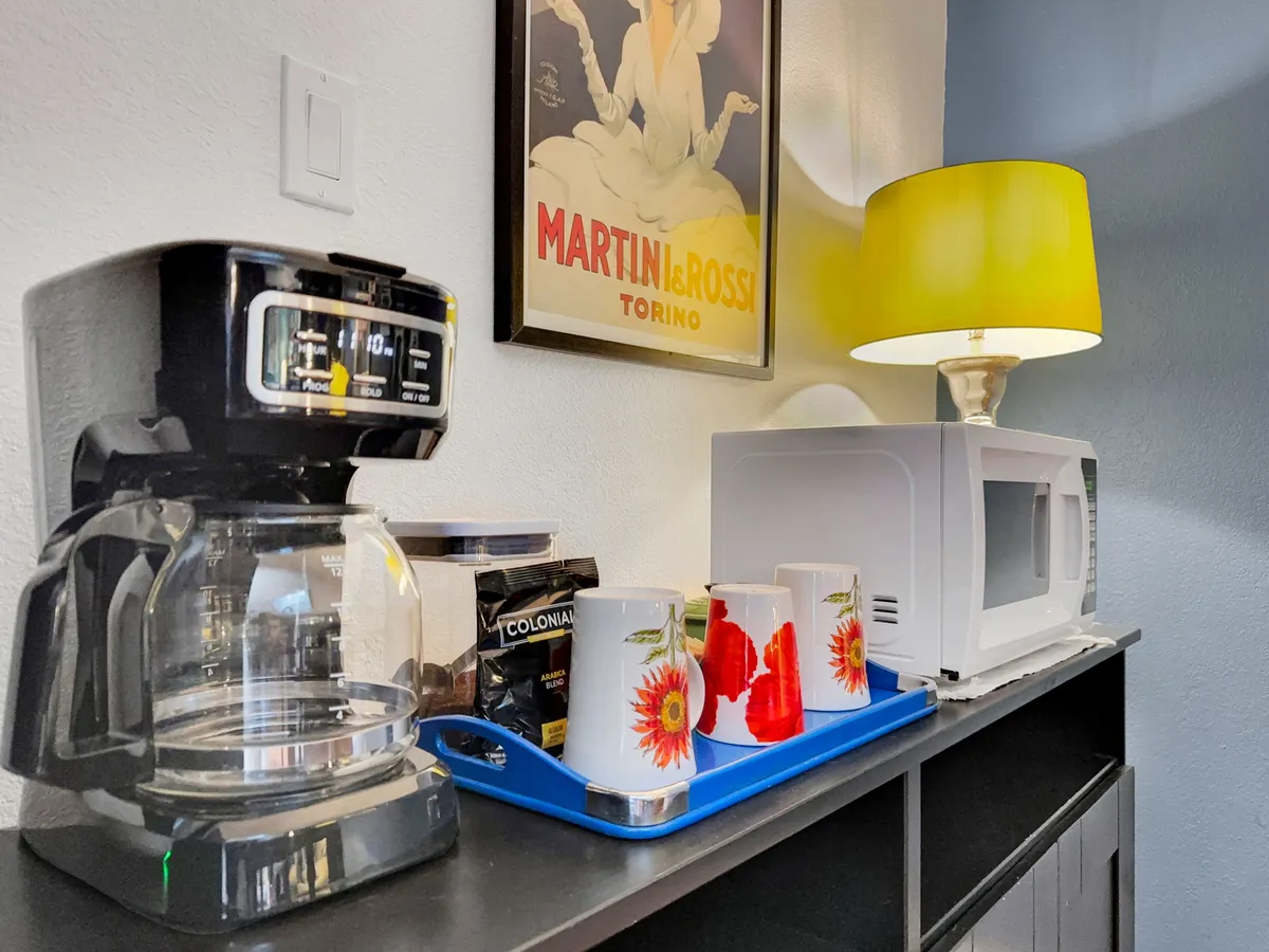 A coffee maker sits on the counter, surrounded by colorful mugs, a yellow lamp, a retro poster, and a microwave. The cheerful kitchen setup suggests a morning routine, inviting guests to start their day with a fresh cup before heading out.