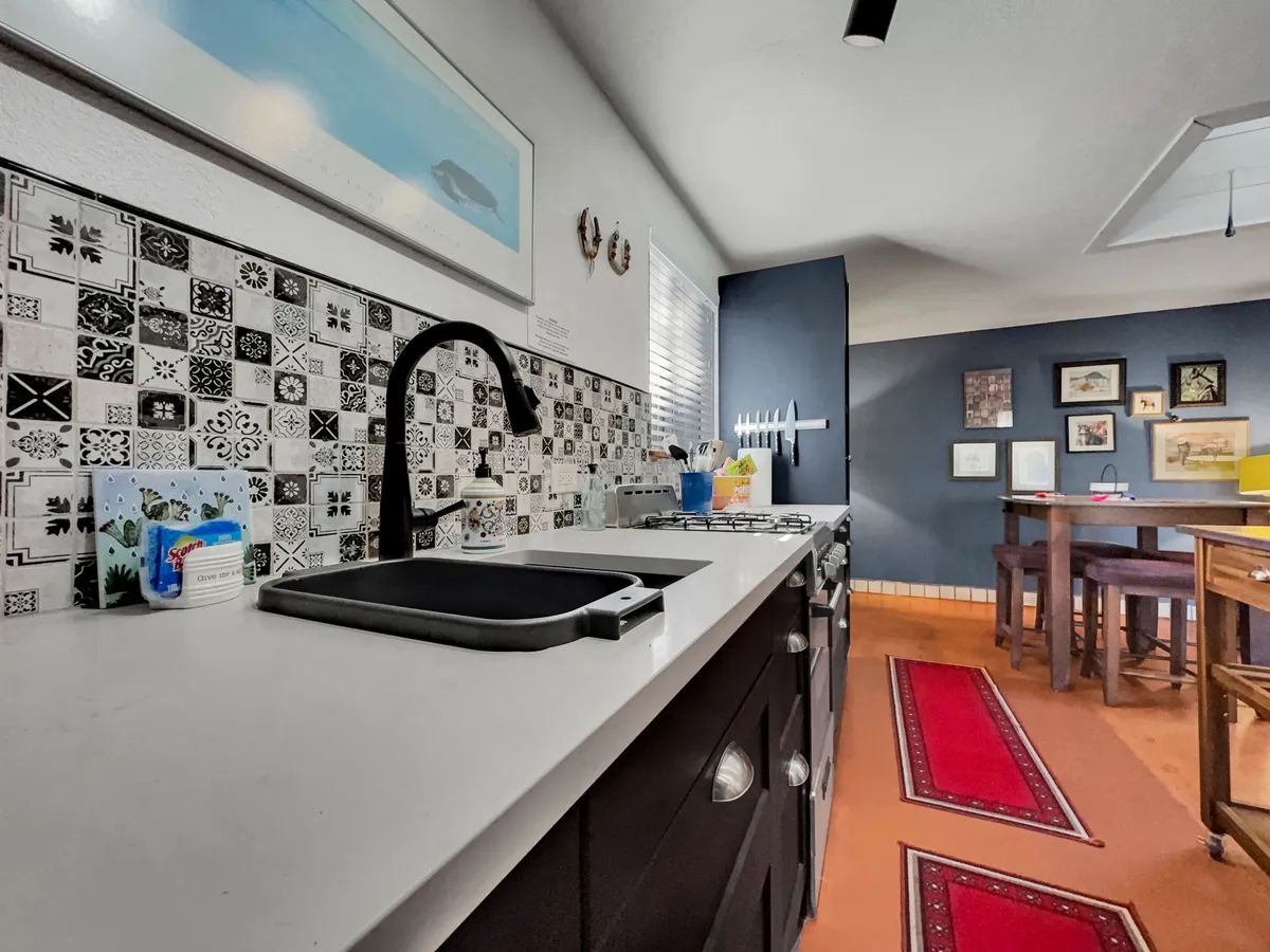 A vibrant kitchen with a decorative tile backsplash, modern sink, colorful artwork, and a view into the adjoining dining area. The decor is lively and the appliances are modern, visually connecting the cooking area to dining and social spaces.