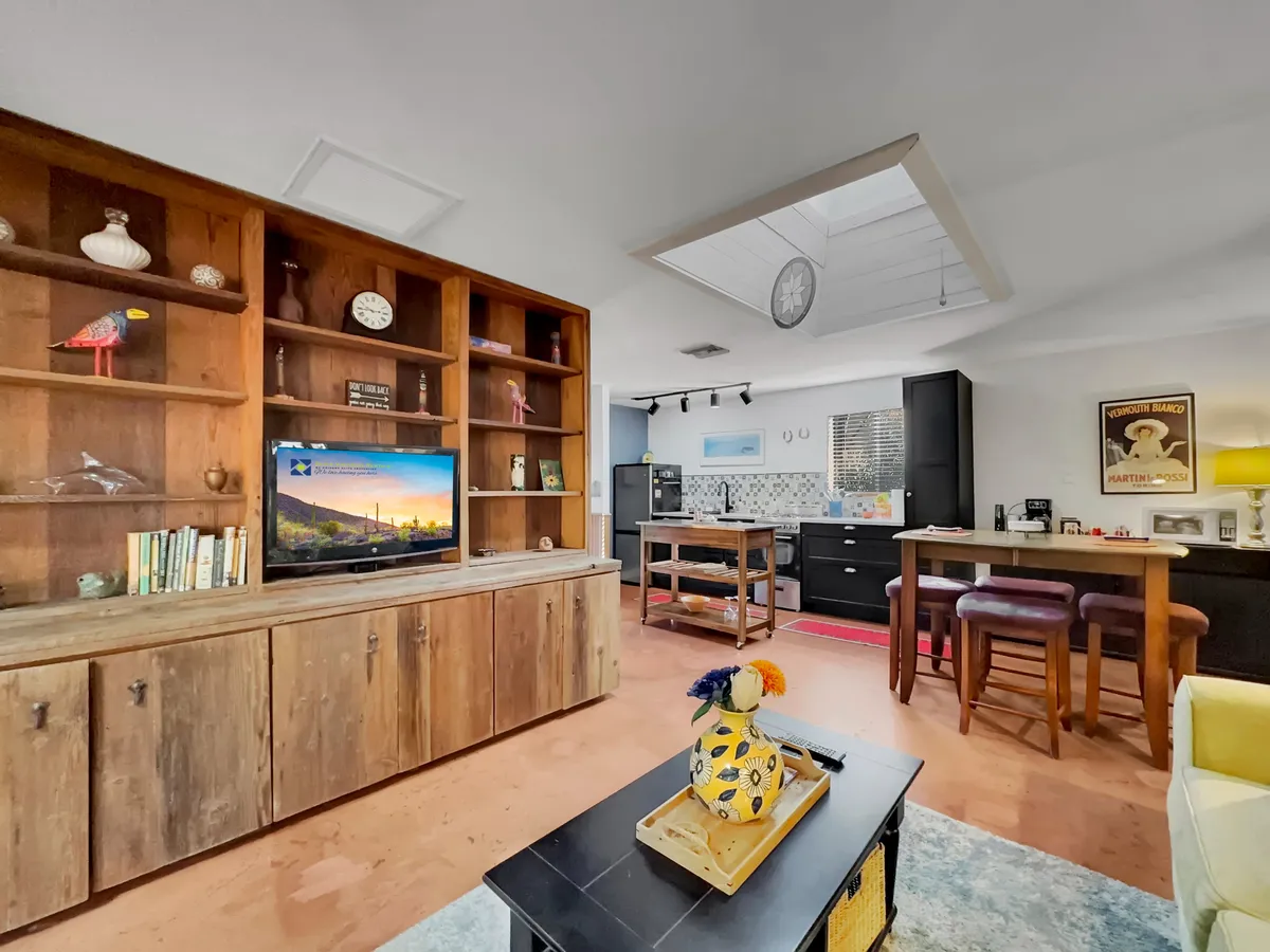 A warm, inviting interior featuring a bar stool dining area adjacent to wooden shelves adorned with decorative items. The television displays a scenic image, while a colorful vase sits on the coffee table. The scene highlights a casual, social space where meals can be enjoyed or prepared.