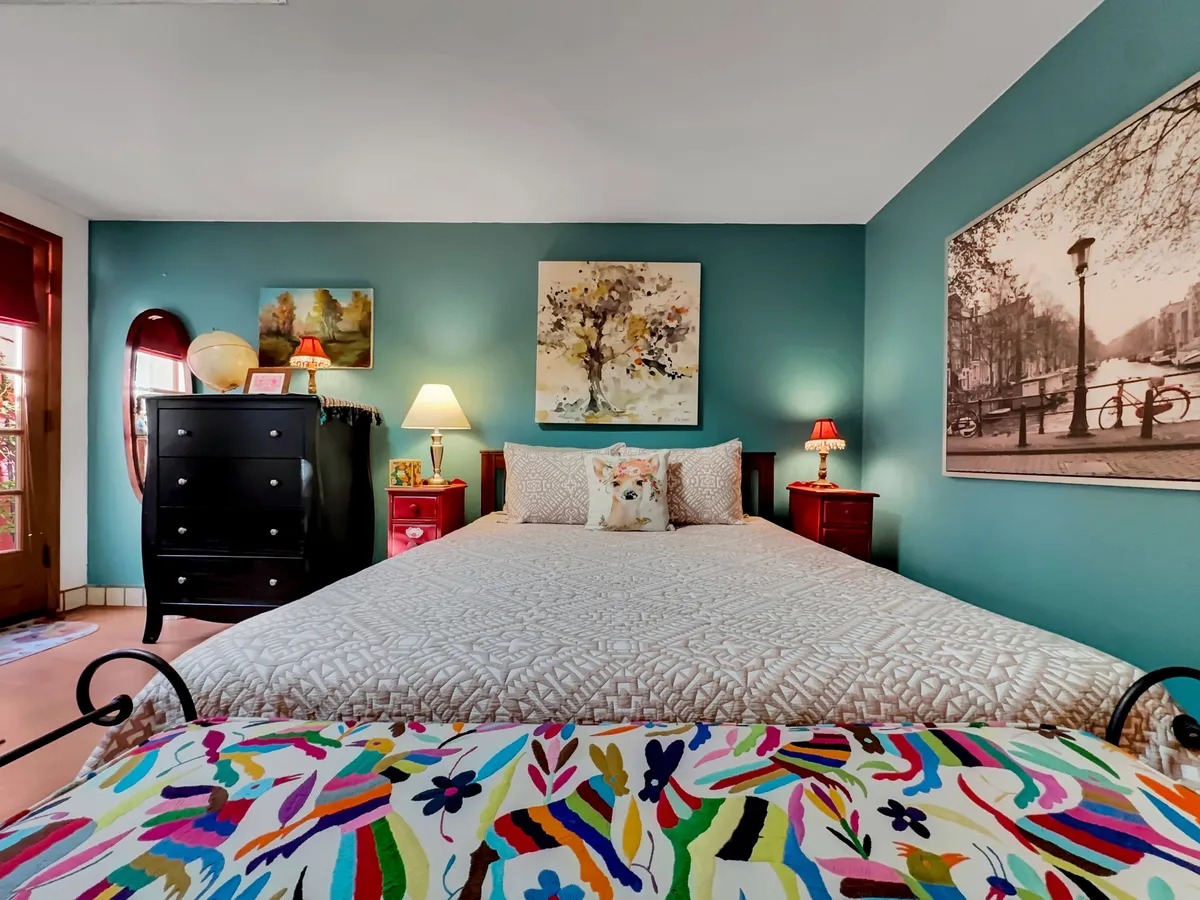 A cheerful bedroom with a colorful bedspread, bedside lamps, art-adorned walls, and a black dresser. The overall feel is warm and comforting, ideal for restful sleep and relaxation.