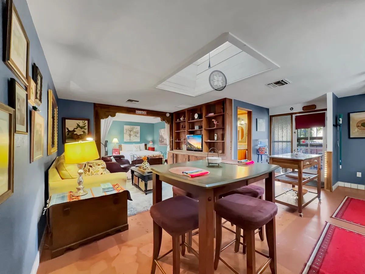A cozy and lively interior with a yellow sofa, a dining table set for meals, decorative wall art, and a bookshelf with a TV. The room exudes a sense of family gatherings and friendly get-togethers, making it ideal for enjoying home-cooked dishes after shopping at local markets.
